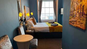 A hotel room with a bed, a round table, a chair, a large window, and a painting on the wall.