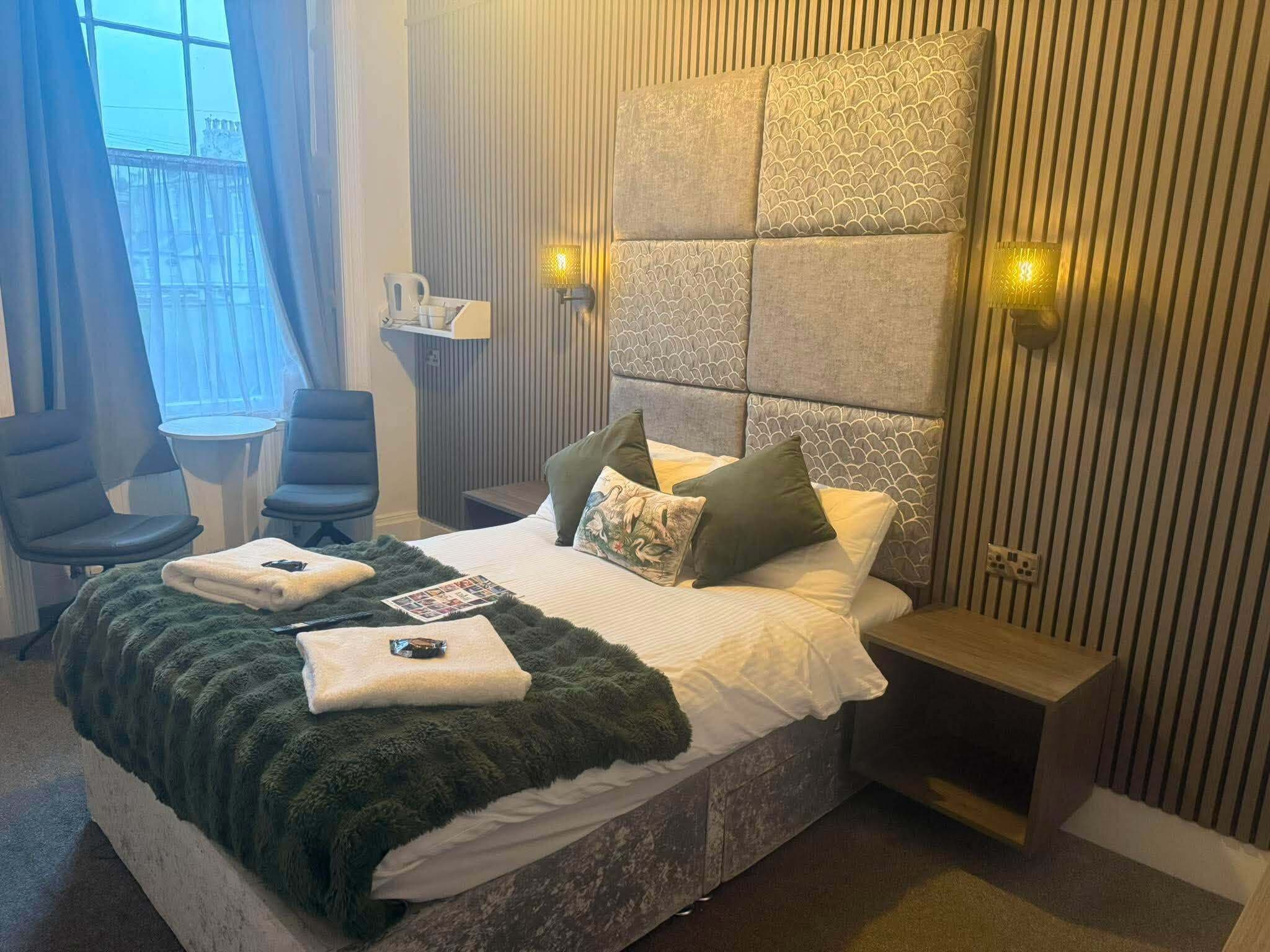 A hotel room with a bed, two chairs, a small table, and a bedside table with a lamp.