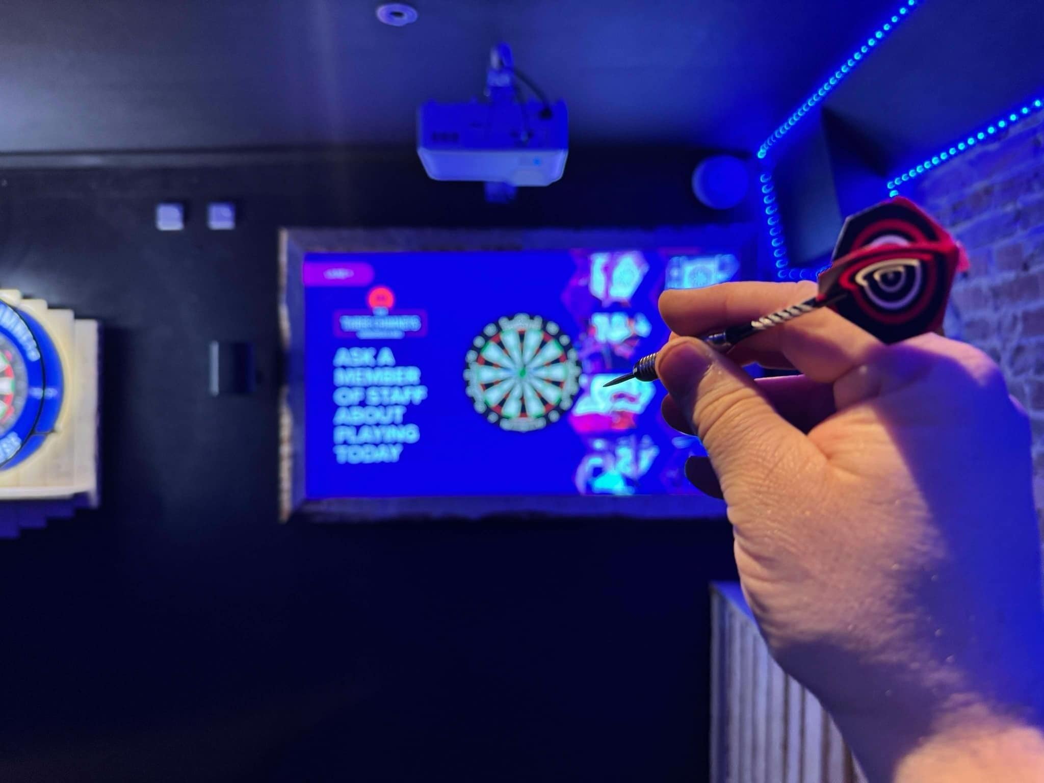 Käsi, jossa on punamusta nuoli, dart-taulu ja näyttö, jossa näkyy pelin käyttöliittymä teksteineen ja grafiikoineen.