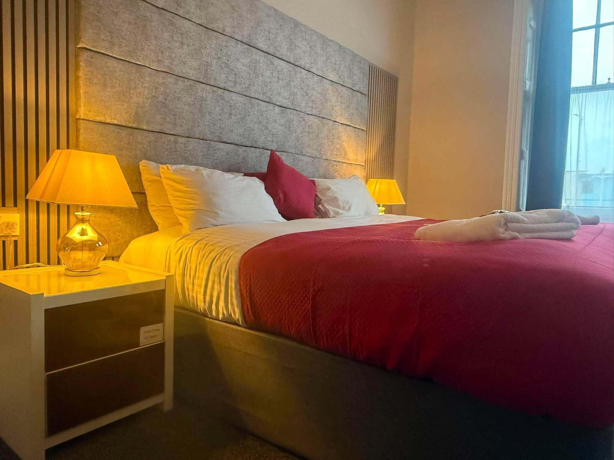 Quarto de hotel com uma cama, duas luminárias de cabeceira, uma mesinha de cabeceira e uma janela.