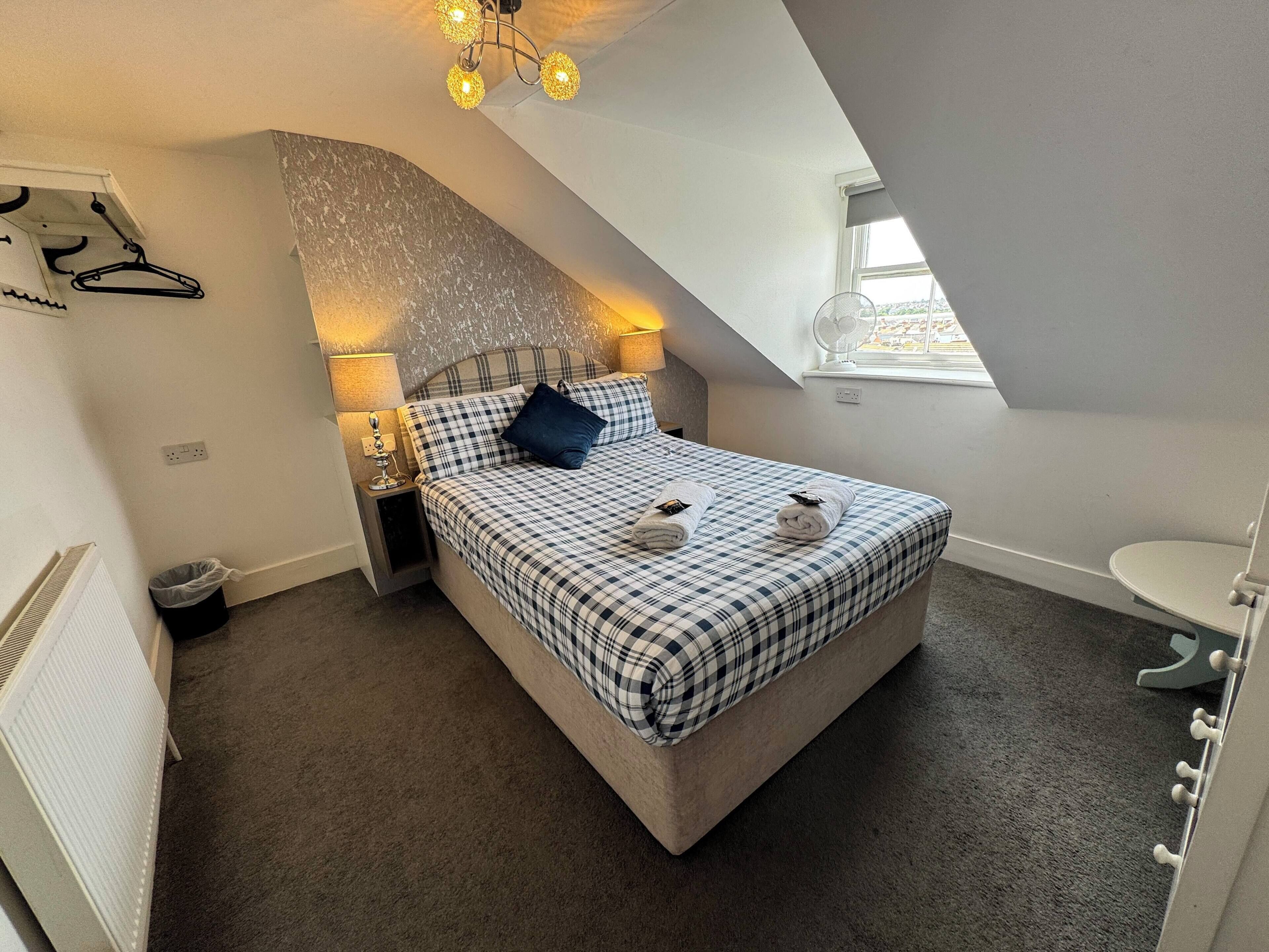 A bedroom with a checkered bedspread, a single pillow, two rolled towels, a bedside lamp, a hanging coat rack, and a window with a view.