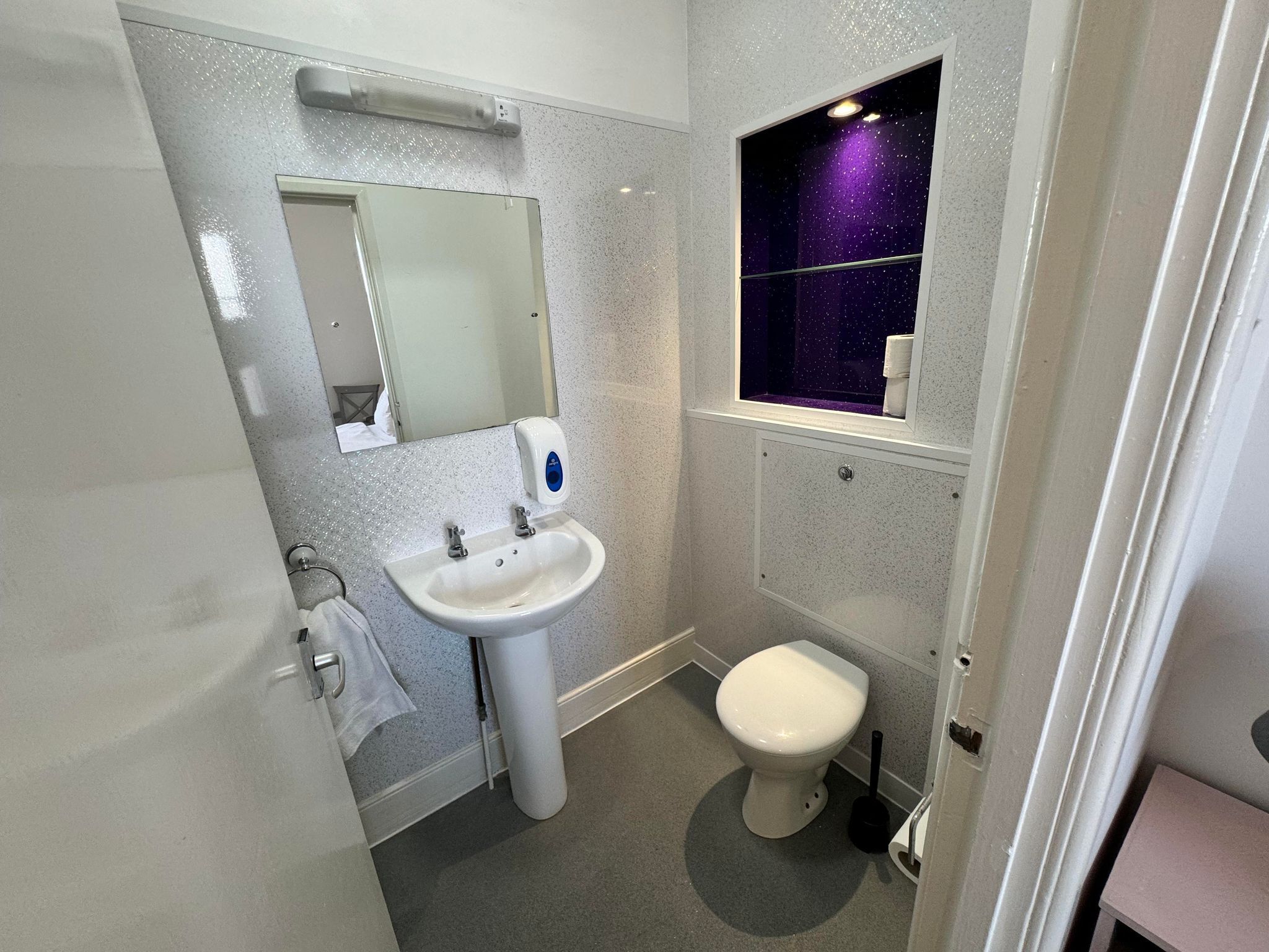 A compact bathroom with a white sink, a mirror, a toilet, and a window with a purple starry view.