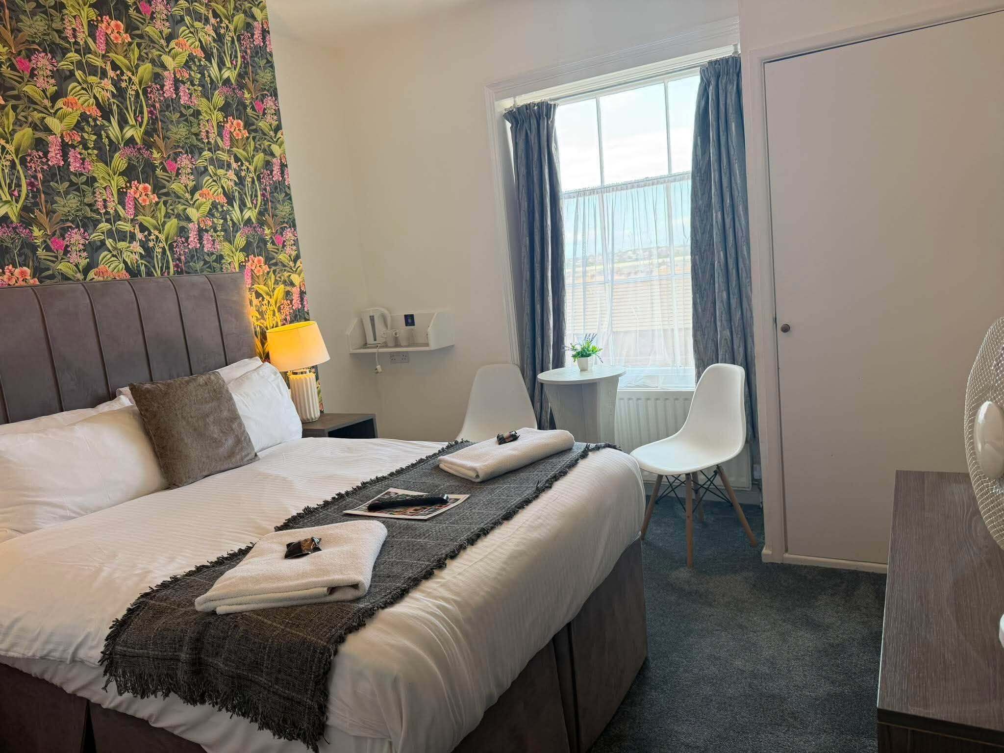 A hotel room with a bed, a desk, a chair, a window with curtains, and a wall with a floral pattern.