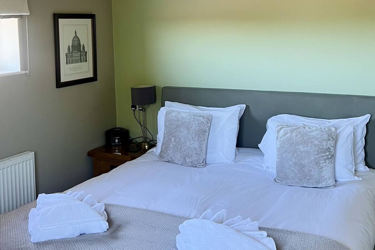 A neatly made double bed with white linens and two decorative pillows, a bedside table with a lamp, and a framed picture on the wall.