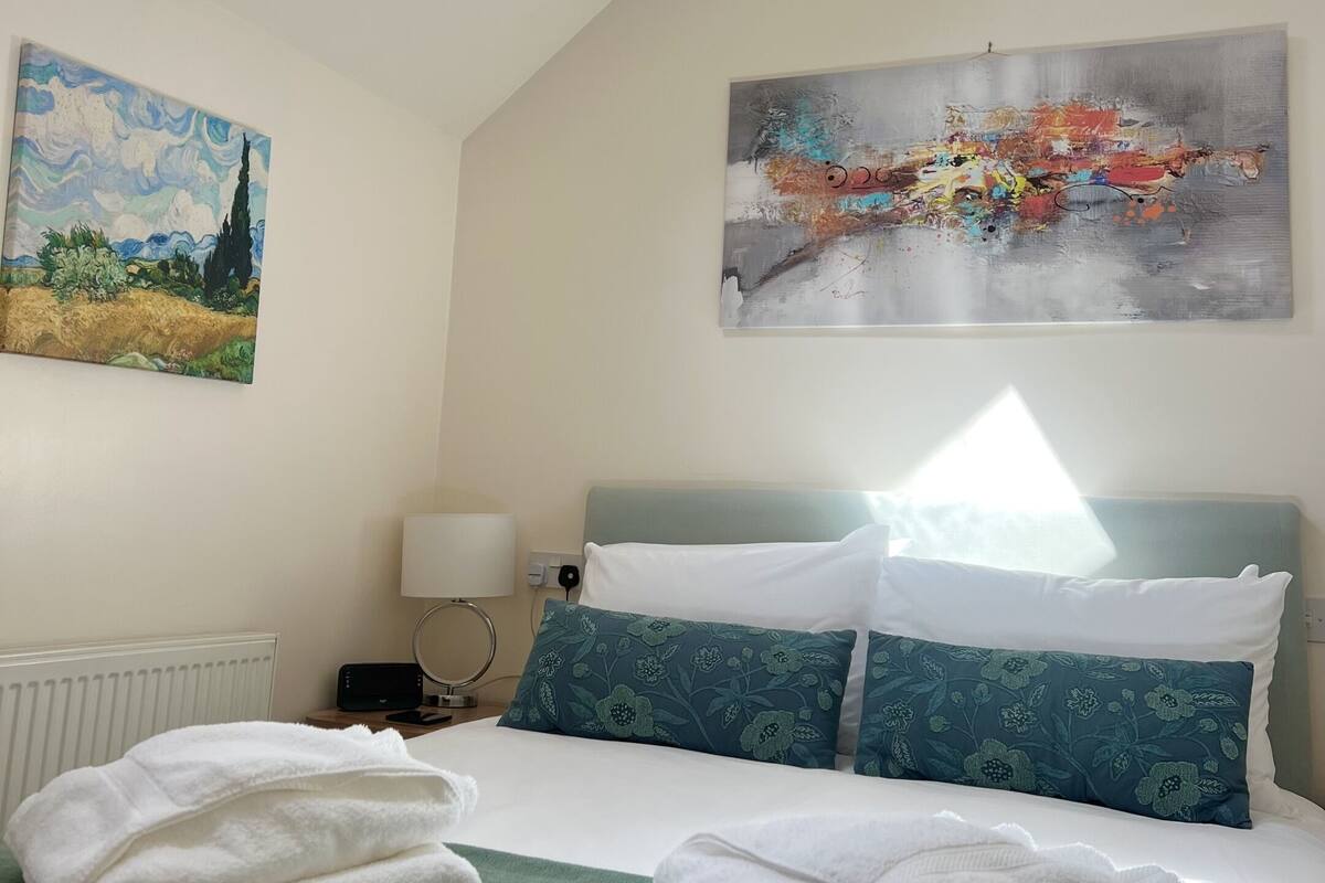 A bedroom with a skylight, a bed with white and blue pillows, a painting of a landscape, and a modern abstract painting.