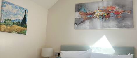 A bedroom with a skylight, a bed with white and blue pillows, a painting of a landscape, and a modern abstract painting.