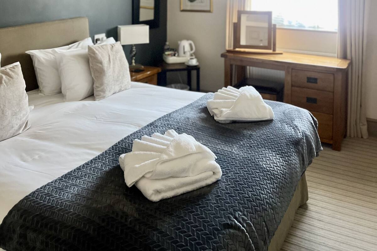 A hotel room with a bed, a desk, a chair, a window with curtains, and a picture on the wall.