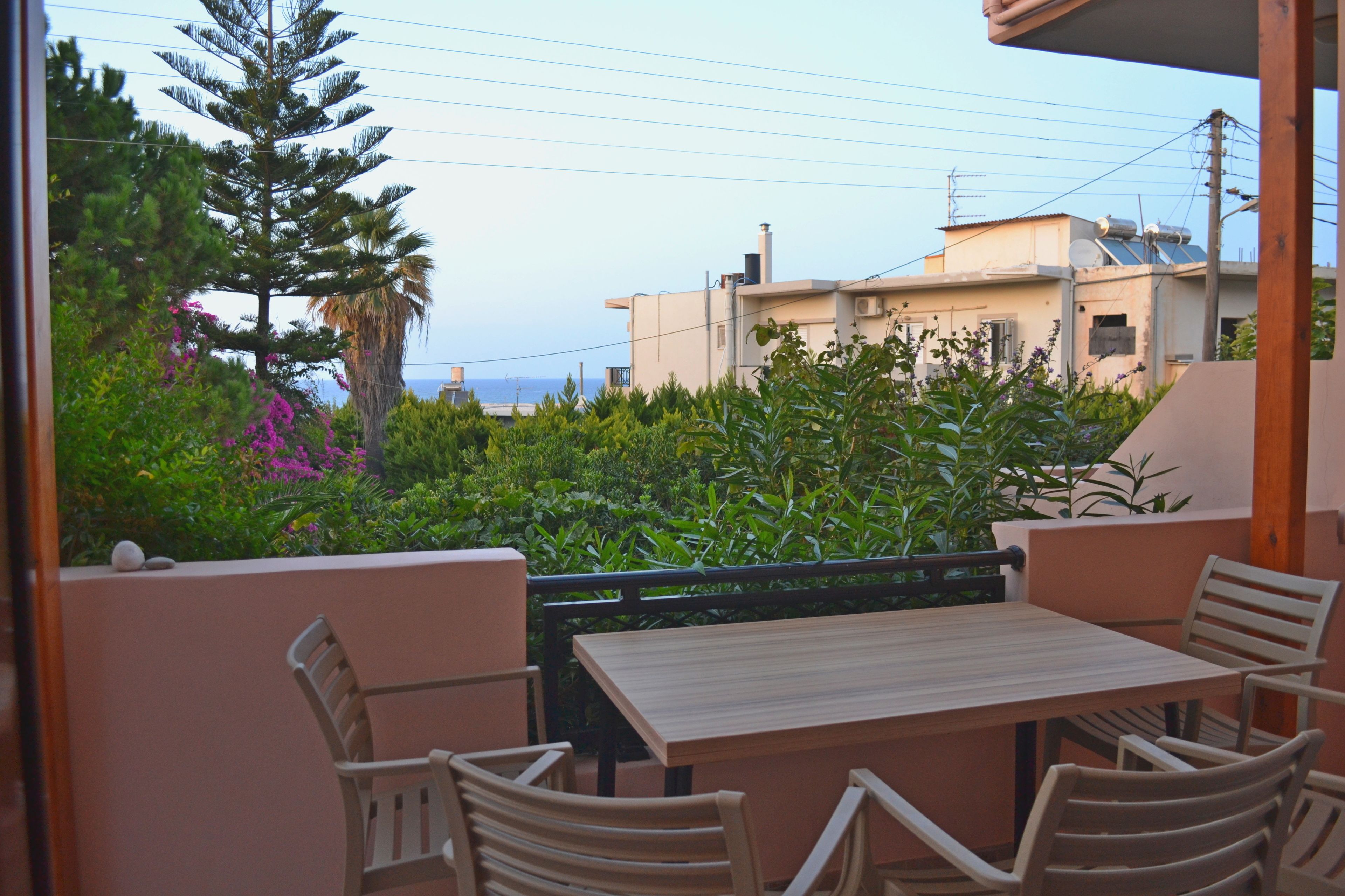 A balcony with a wooden table and chairs, surrounded by lush greenery and a view of buildings and the sea.