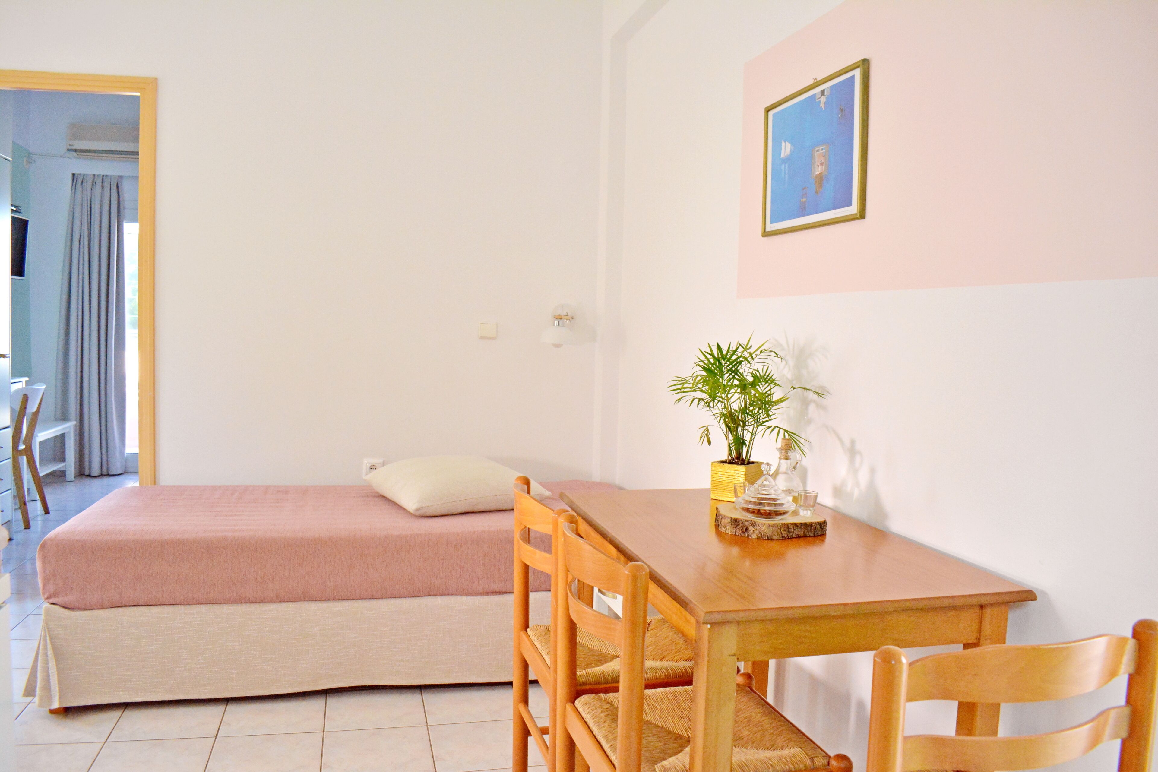 Um quarto com uma cama, uma mesa de madeira com cadeiras, um vaso de plantas e um quadro na parede.