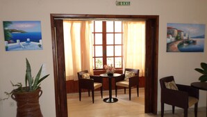 A room with a wooden floor, a round table with chairs, a large window with curtains, and paintings on the walls. - Tina Economy Hotel (Chania)
