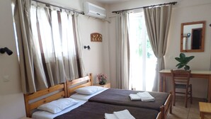 A double bed with two pillows, a wooden nightstand, a desk with a chair, and a potted plant. - Tina Economy Hotel (Chania)