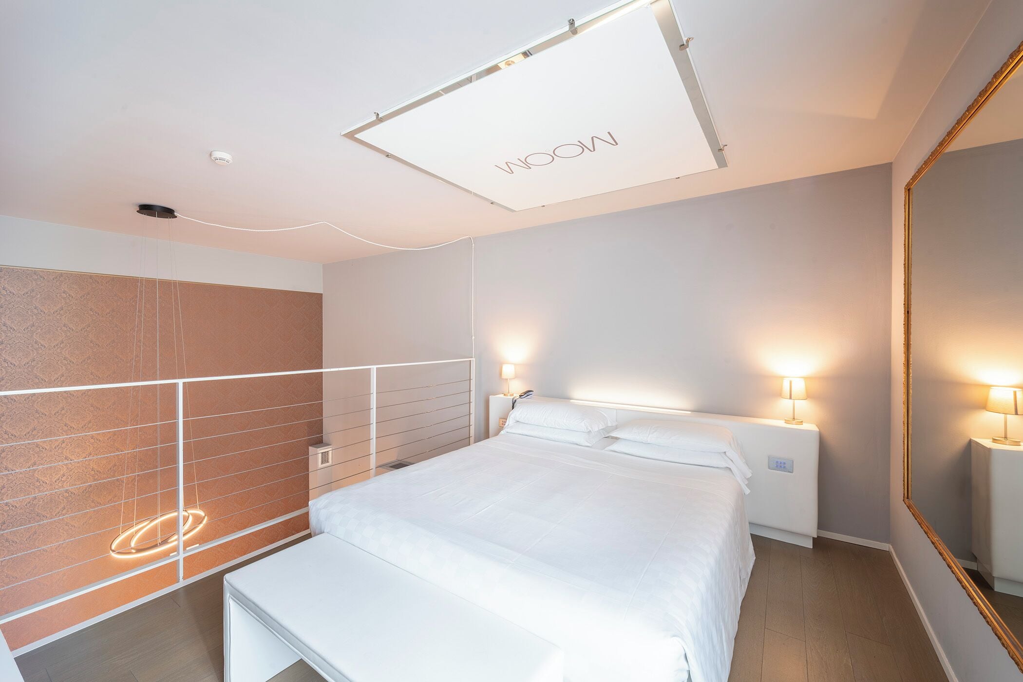 A modern hotel room with a large bed, bedside lamps, a mirror, and a ceiling light fixture.