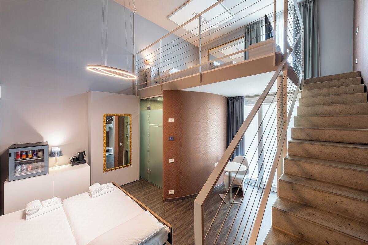 A modern hotel room with a bed, a minibar, a mirror, a staircase, and a view of the city.