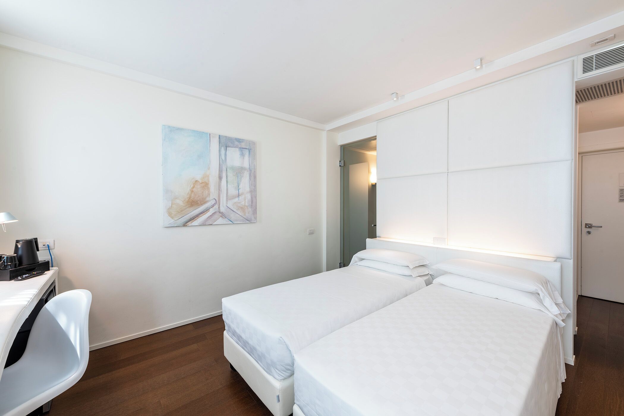 A hotel room with two single beds, a desk with a chair, a painting on the wall, and a door leading to another room.