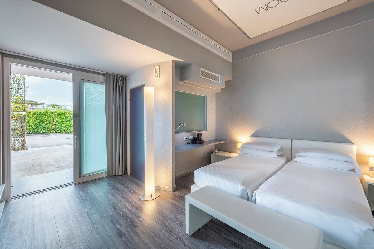 A modern hotel room with two beds, a large window, a sliding door, a bedside table, and a built-in shelf.