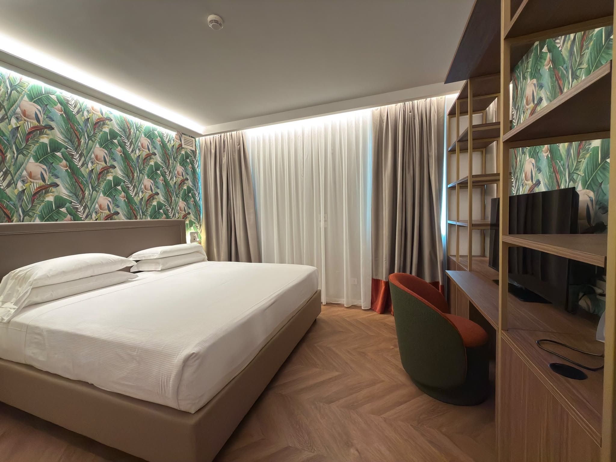 Habitación de hotel con cama, cortinas, televisor y pared con estampado tropical.