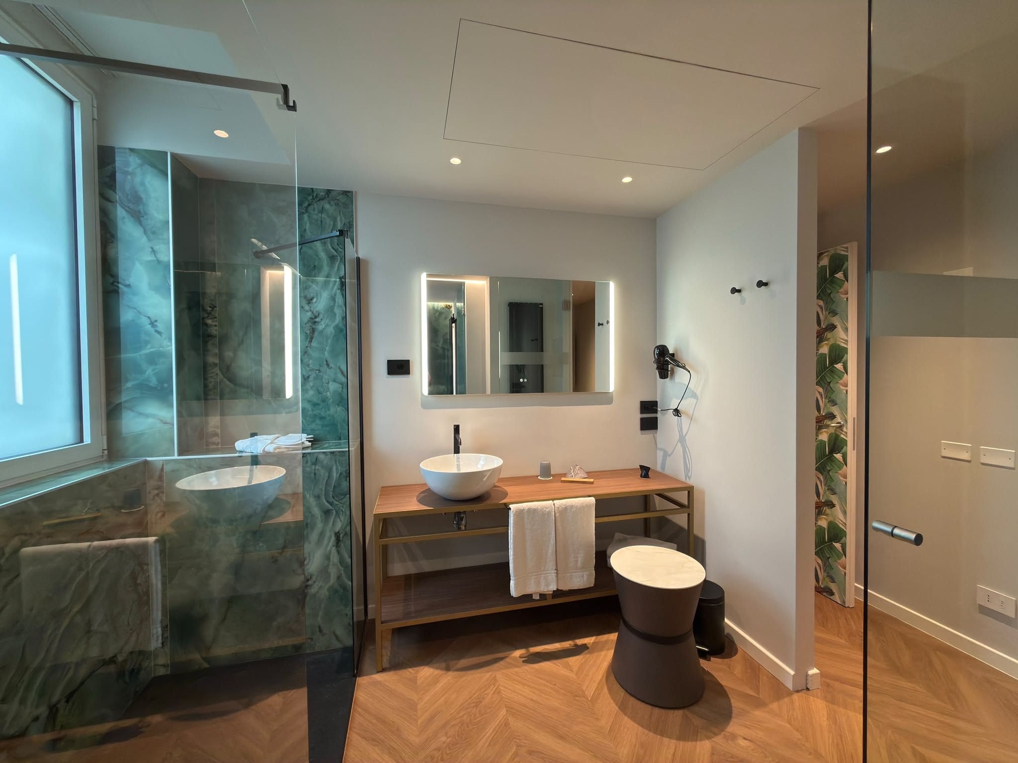 Un baño moderno con ducha envuelta en vidrio, un espejo grande, un tocador de madera y un taburete de madera redondo.