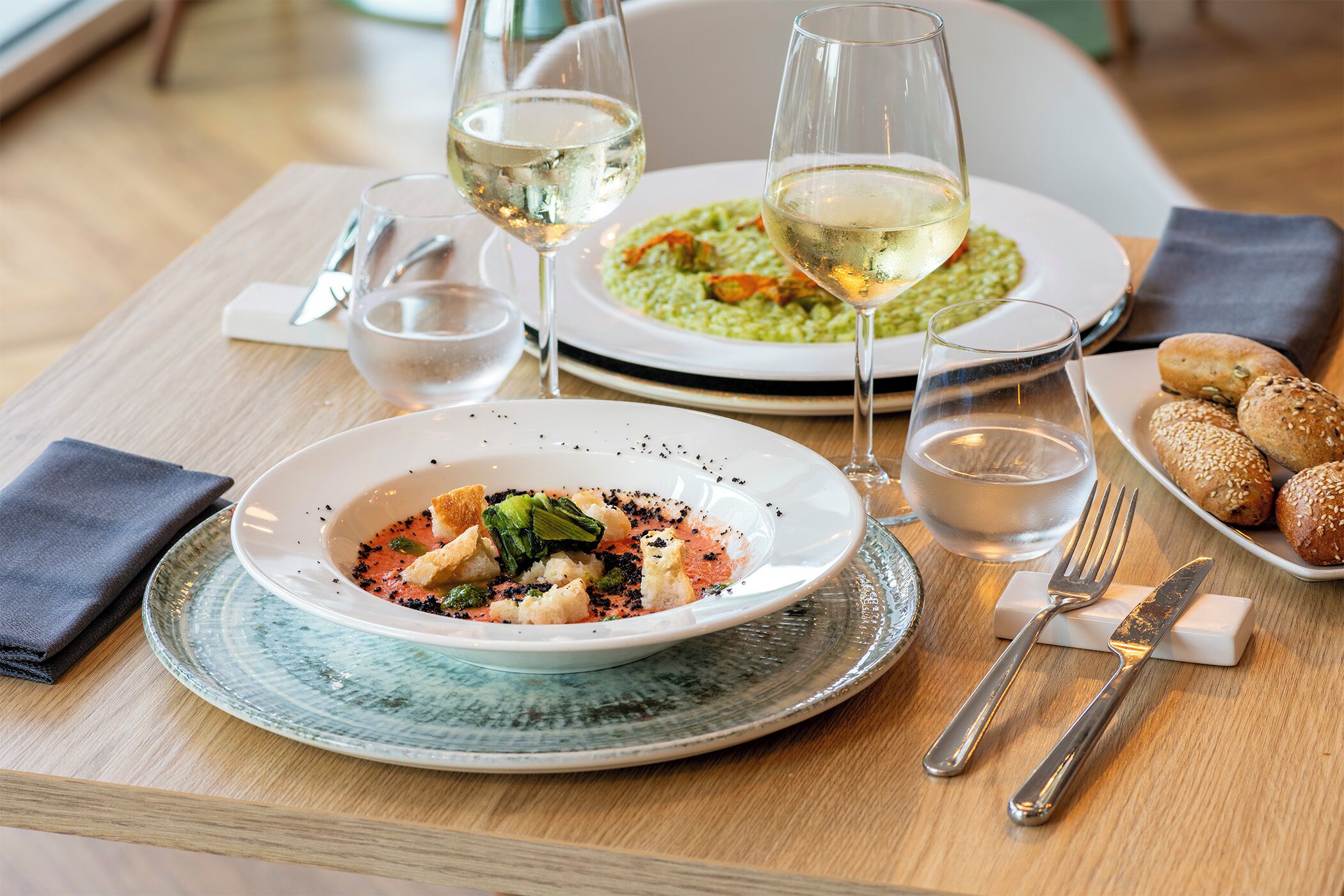 Una mesa de comedor con platos de comida, copas de vino blanco y cubiertos.