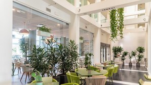 Un espacio interior moderno con varias mesas y sillas, rodeado de plantas en macetas y con un muro de vidrio.