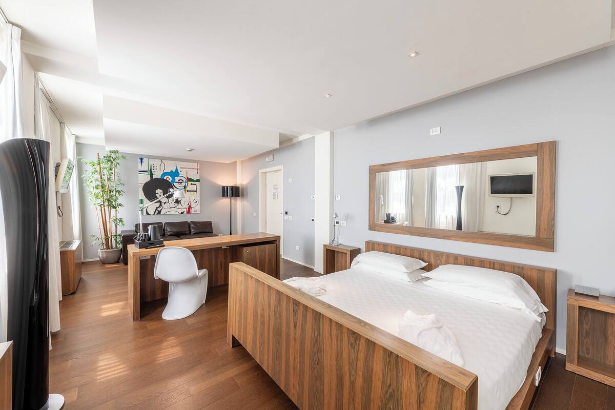 A modern hotel room with a wooden bed, a desk, and a chair. There is a large mirror on the wall and a TV mounted above it.