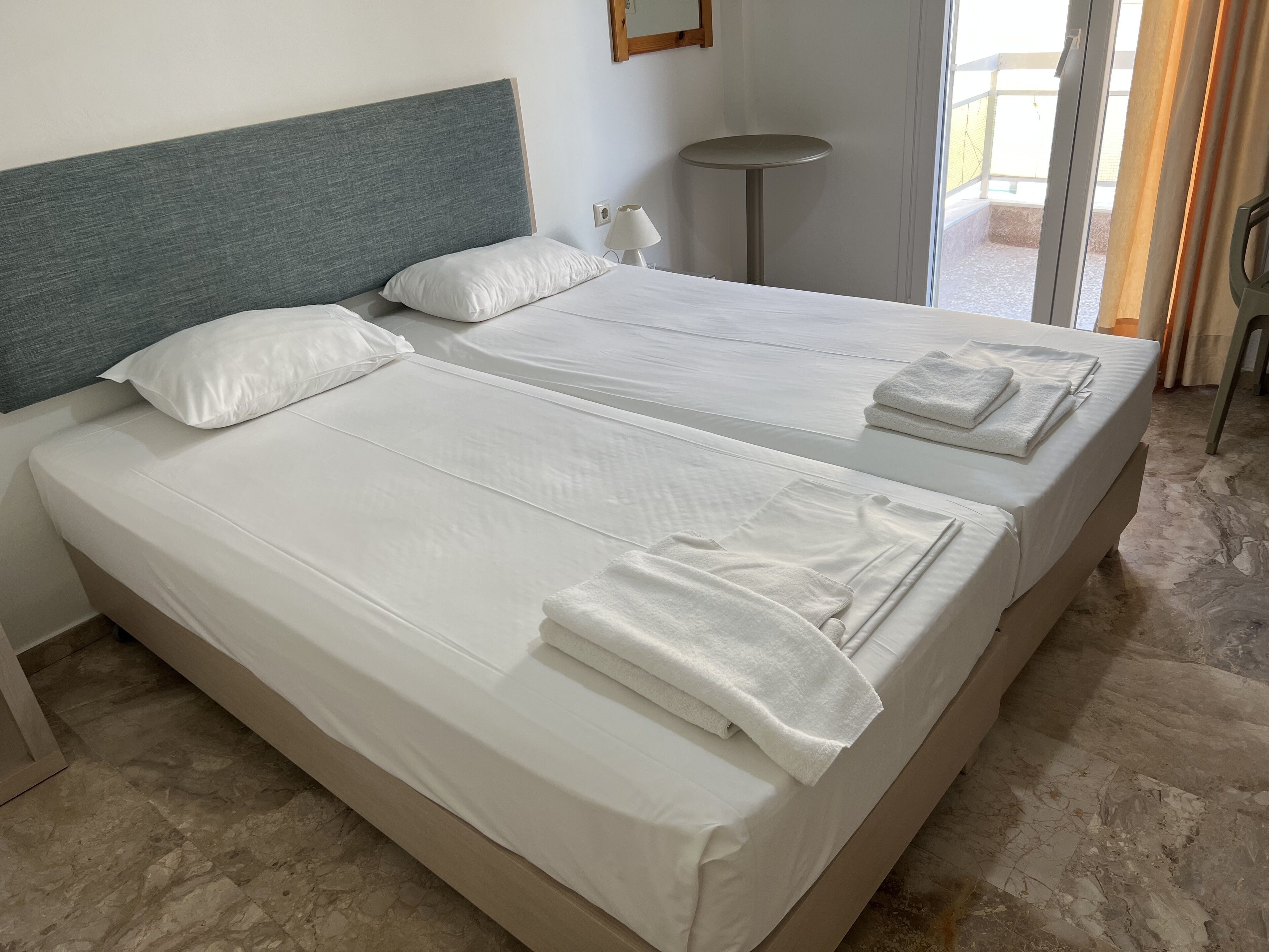 Camera d'albergo con due letti, ognuno con lenzuola e asciugamani bianchi, un tavolino rotondo e vista sull'esterno tramite una porta scorrevole.