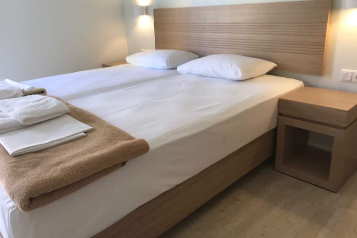 Een net opgemaakt bed met witte lakens en een bruine plaid, een houten hoofdbord en een klein nachtkastje met een lade.
