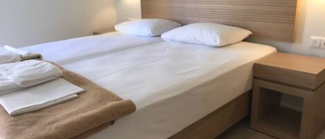 Ein ordentlich bezogenes Bett mit weißen Laken und einer braunen Tagesdecke, ein Holz-Kopfschild und ein kleiner Nachttisch mit Schublade.