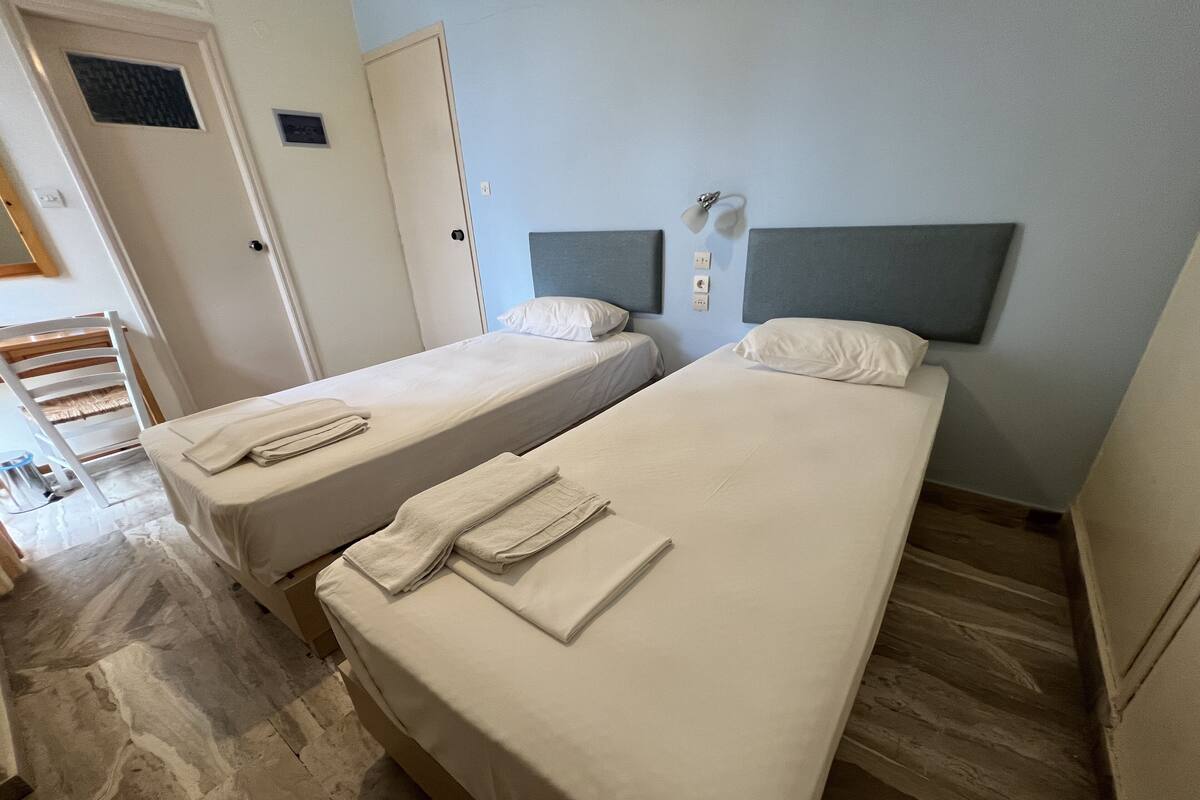 Twee enkele bedden met hoofdborden, een tafeltje en een stoel in een kamer met betegelde vloer.