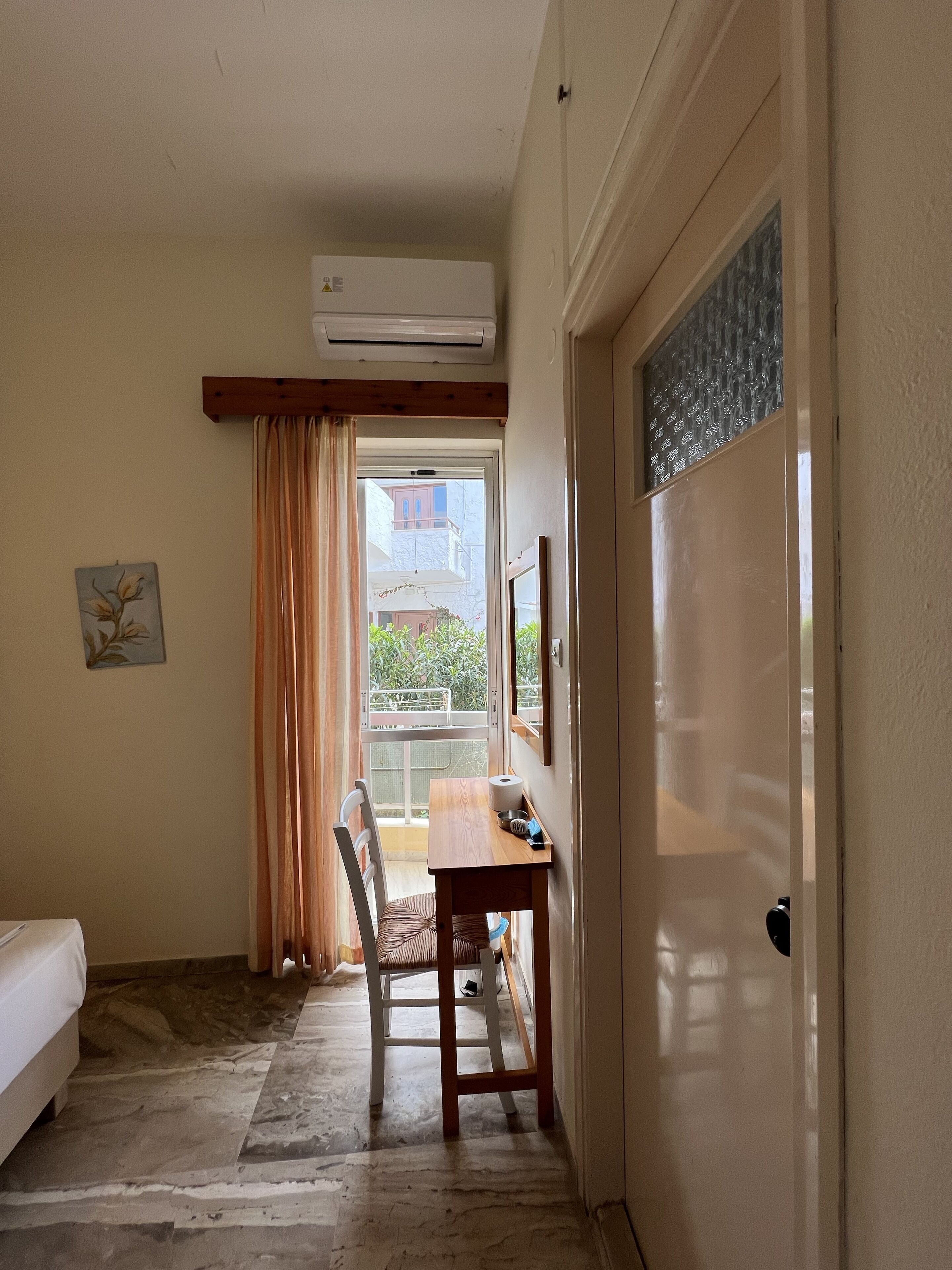 A room with a bed, a wooden table with a chair, an air conditioning unit, a window with curtains, and a view of buildings outside.