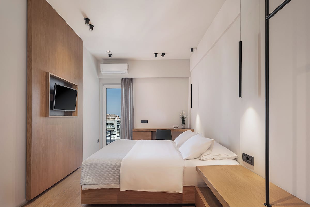 A modern hotel room with a wooden bed, a flat-screen TV mounted on the wall, a small desk with a chair, and a view of the city through a window.