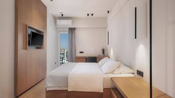 A modern hotel room with a wooden bed, a flat-screen TV mounted on the wall, a small desk with a chair, and a view of the city through a window.