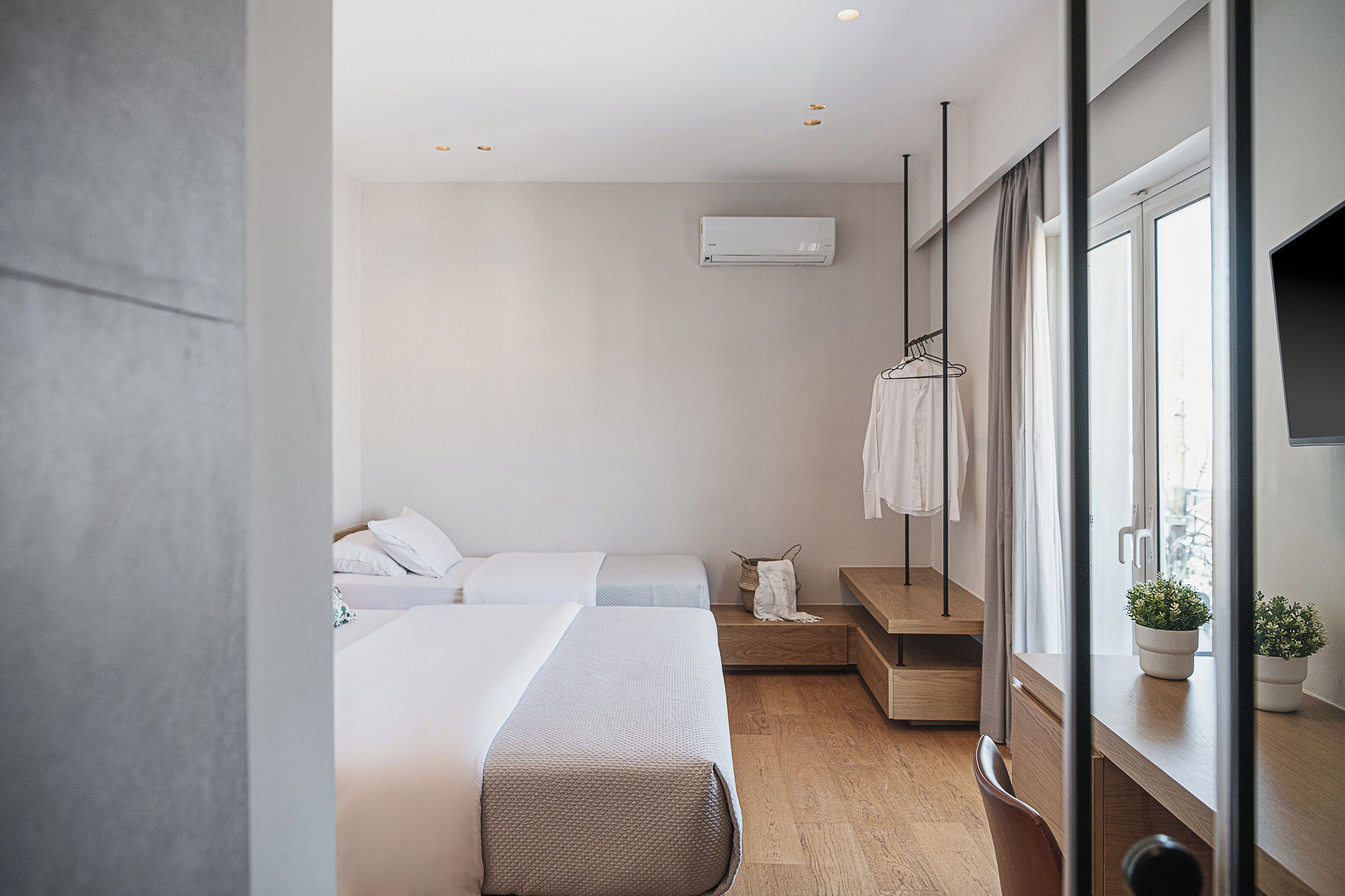 A modern hotel room with two beds, a wooden bench, a small table, a TV, and a window with curtains.