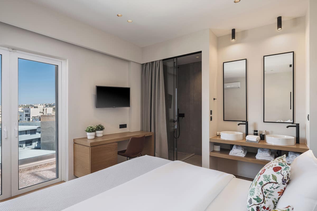 A hotel room with a bed, a desk with a chair, a TV, a shower area, and a bathroom with a sink and mirror.