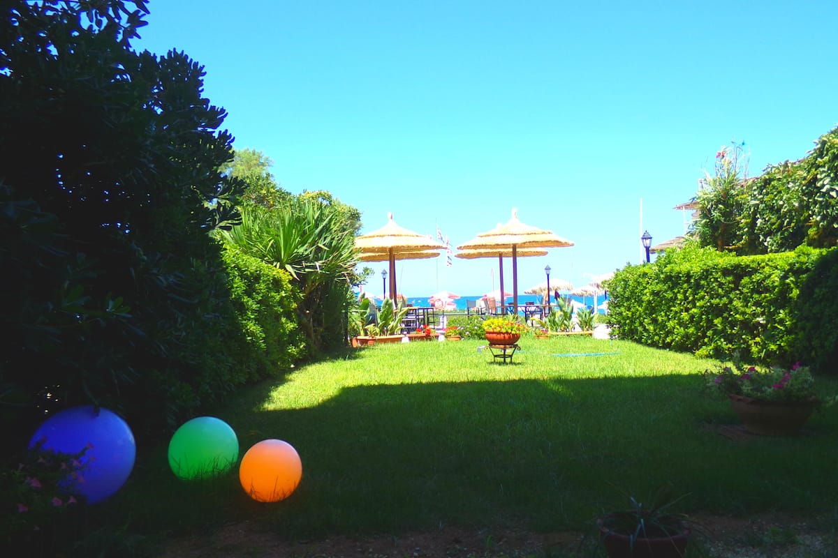 Un jardín con césped, dos sombrillas de paja y una mesa con sillas.