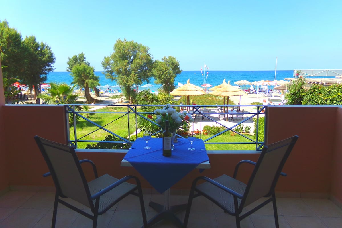 Un balcón con dos sillas y una mesa, con vista a una playa con sombrillas y un cielo azul despejado.