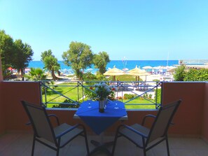 A balcony with two chairs and a table, overlooking a beach with umbrellas and a clear blue sky. - Katerina Seaside Studios (Platanias)