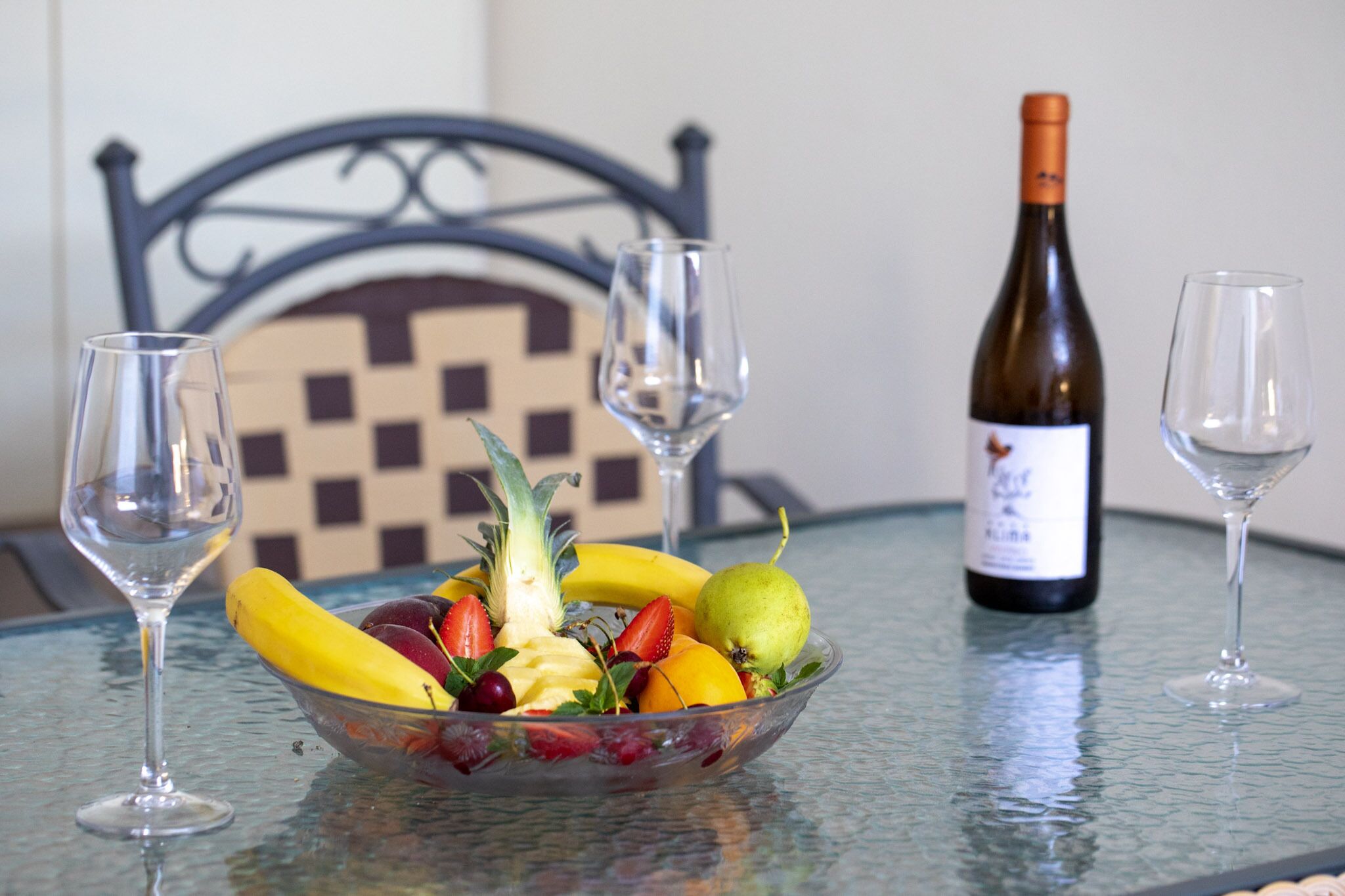 A glass table with a fruit bowl, two wine glasses, and a wine bottle.
