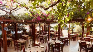Un restaurant en plein air avec des tables et des chaises en bois, abrité par une pergola couverte de feuilles vertes et de fleurs roses.