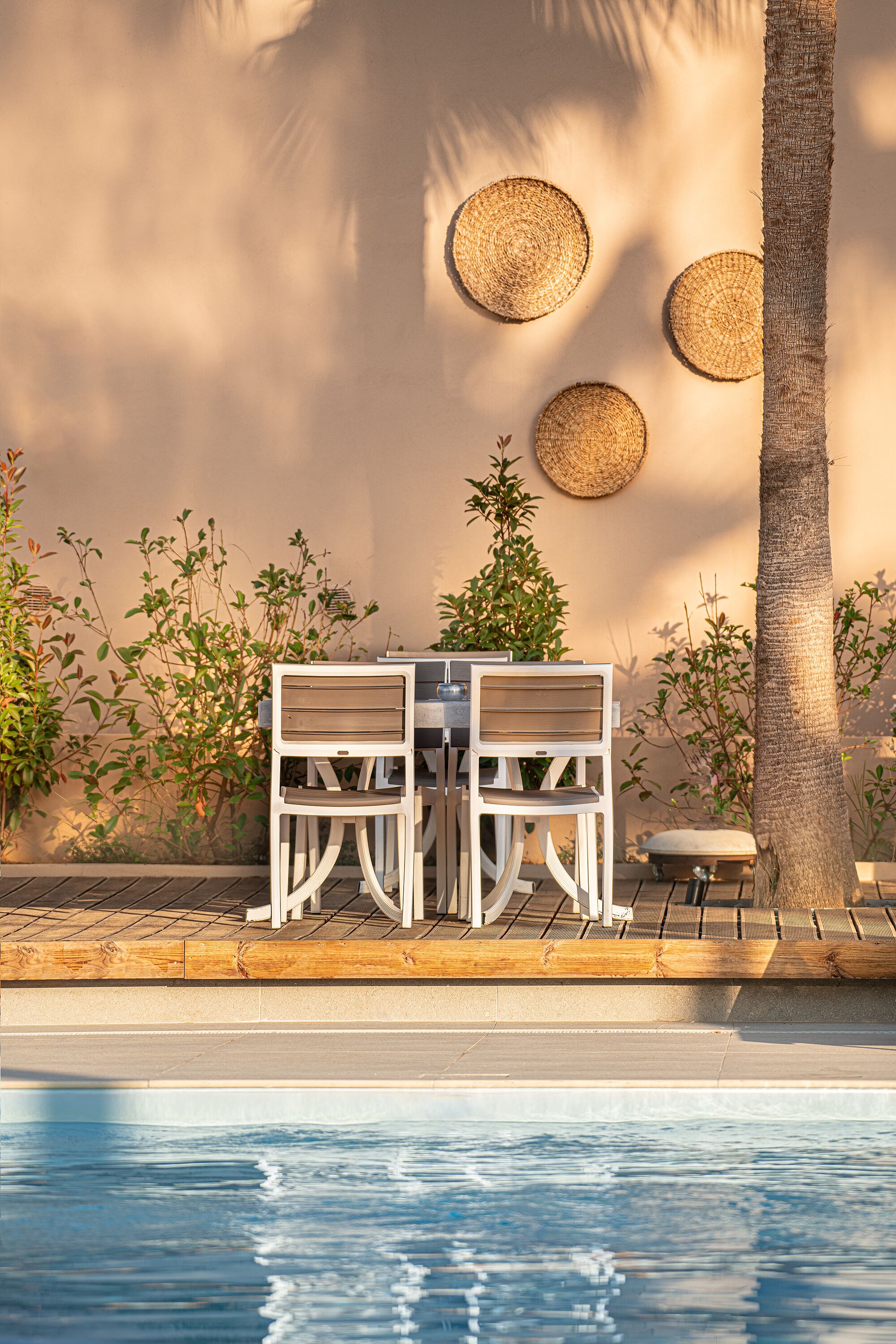 Zona junto a la piscina con una mesa, sillas, una palmera y una pared decorada con esteras tejidas.