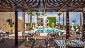 Un espace aménagé au bord de la piscine, avec des chaises longues et des tables, une piscine, et un bâtiment dont le mur arrière est recouvert de verdure.