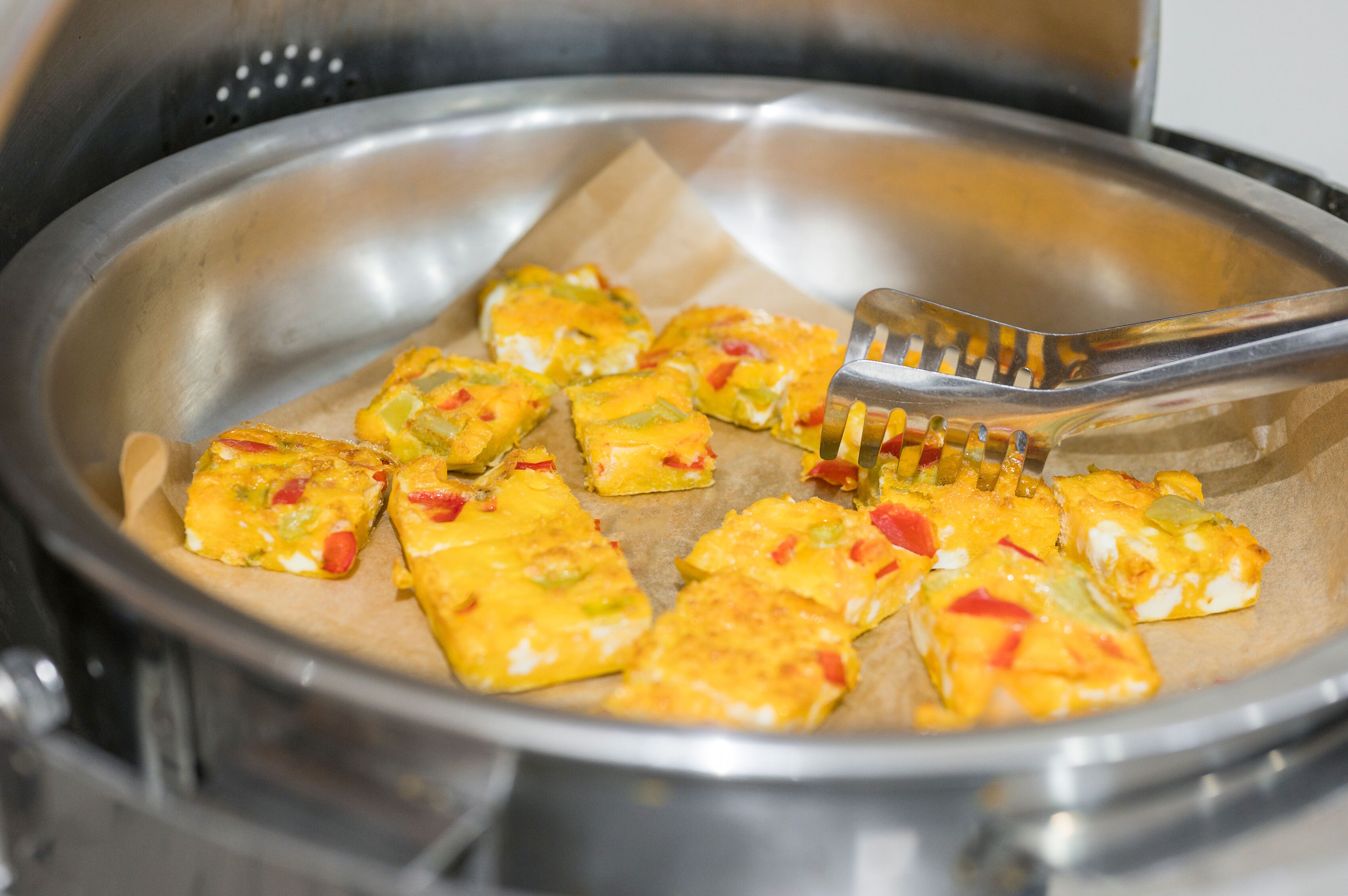 A stainless steel pan with a paper liner containing several pieces of food, possibly a quiche or frittata, being served with a spatula.
