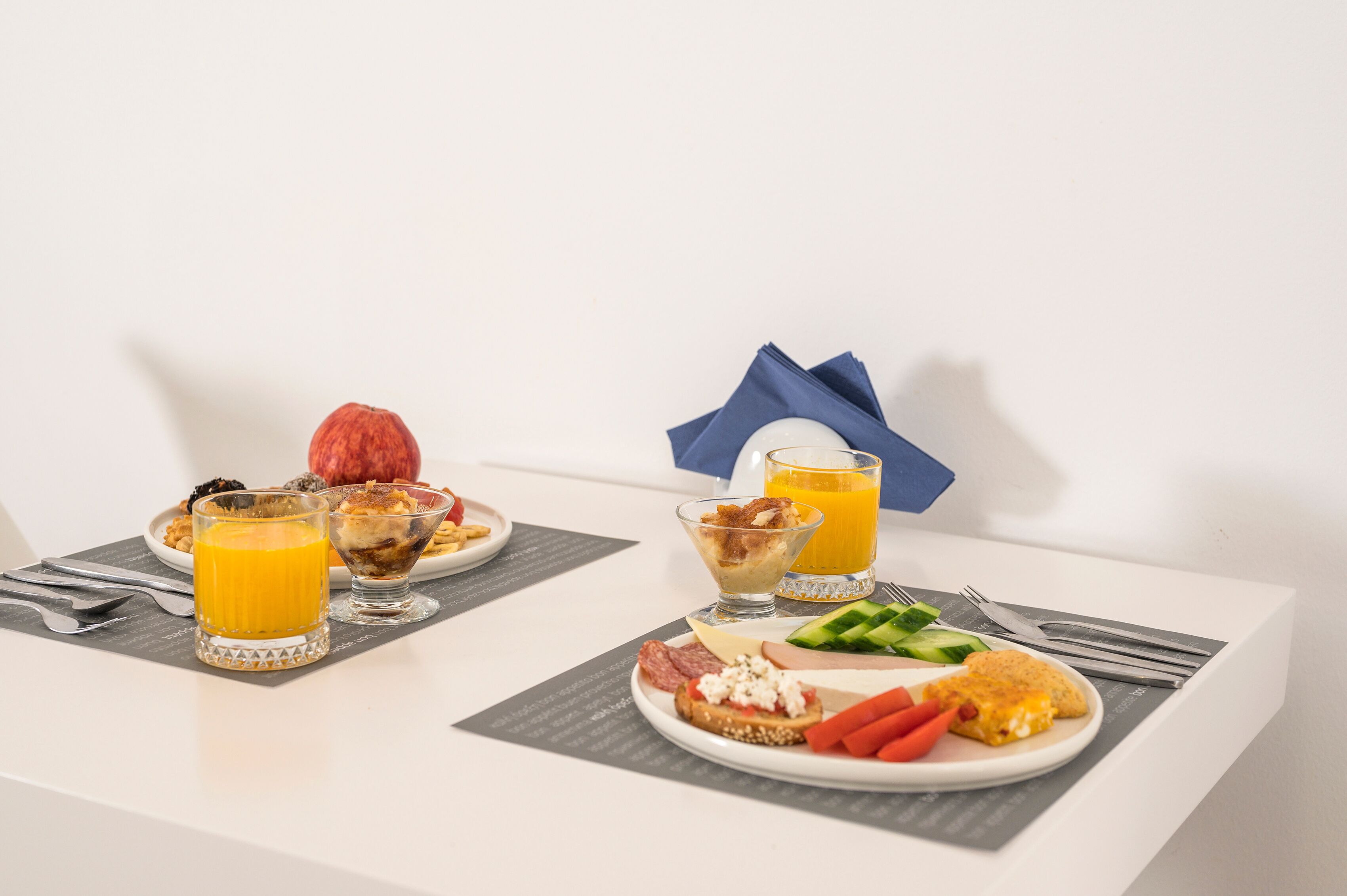 Desayuno servido con dos platos de comida, dos vasos de jugo de naranja y una servilleta azul sobre una mesa blanca.