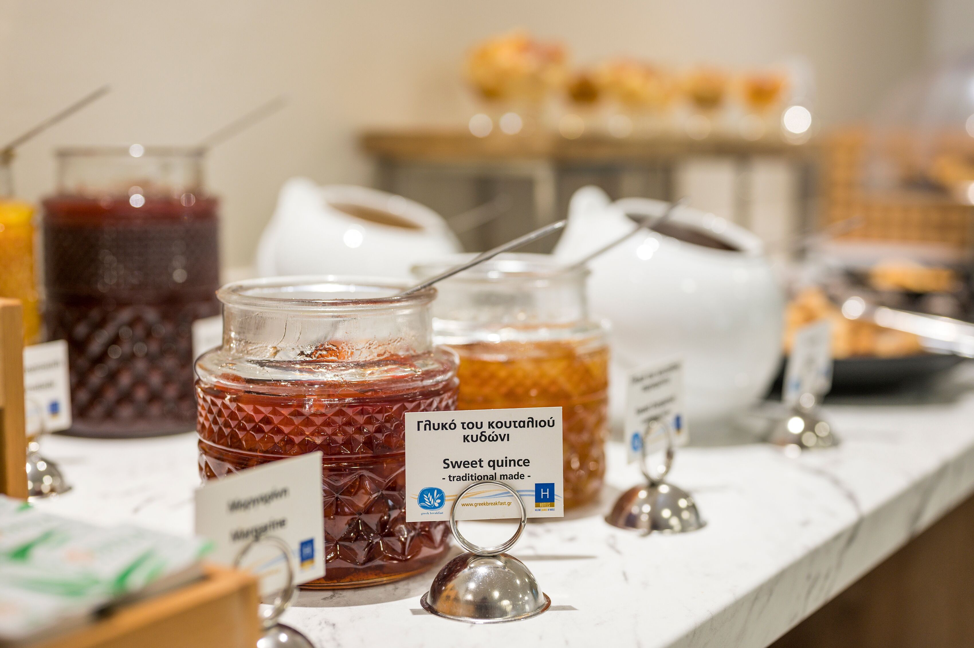 En buffet med forskellige glas med mad, herunder et glas med teksten “Sødsyltet kvæde, traditionel fremstilling.”