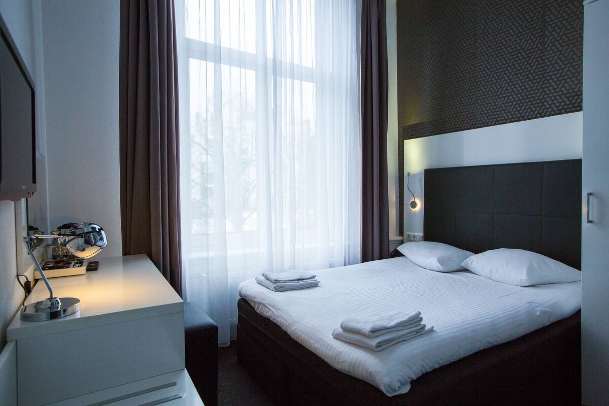 Ett hotellrum med en snyggt bäddad säng, ett nattduksbord med en lampa och ett fönster med tunna, genomskinliga gardiner.