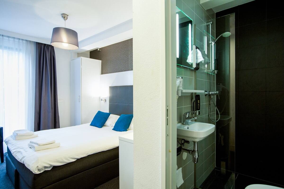 Ett hotellrum med en säng, ett sängbord, en garderob och ett badrum med handfat och dusch.