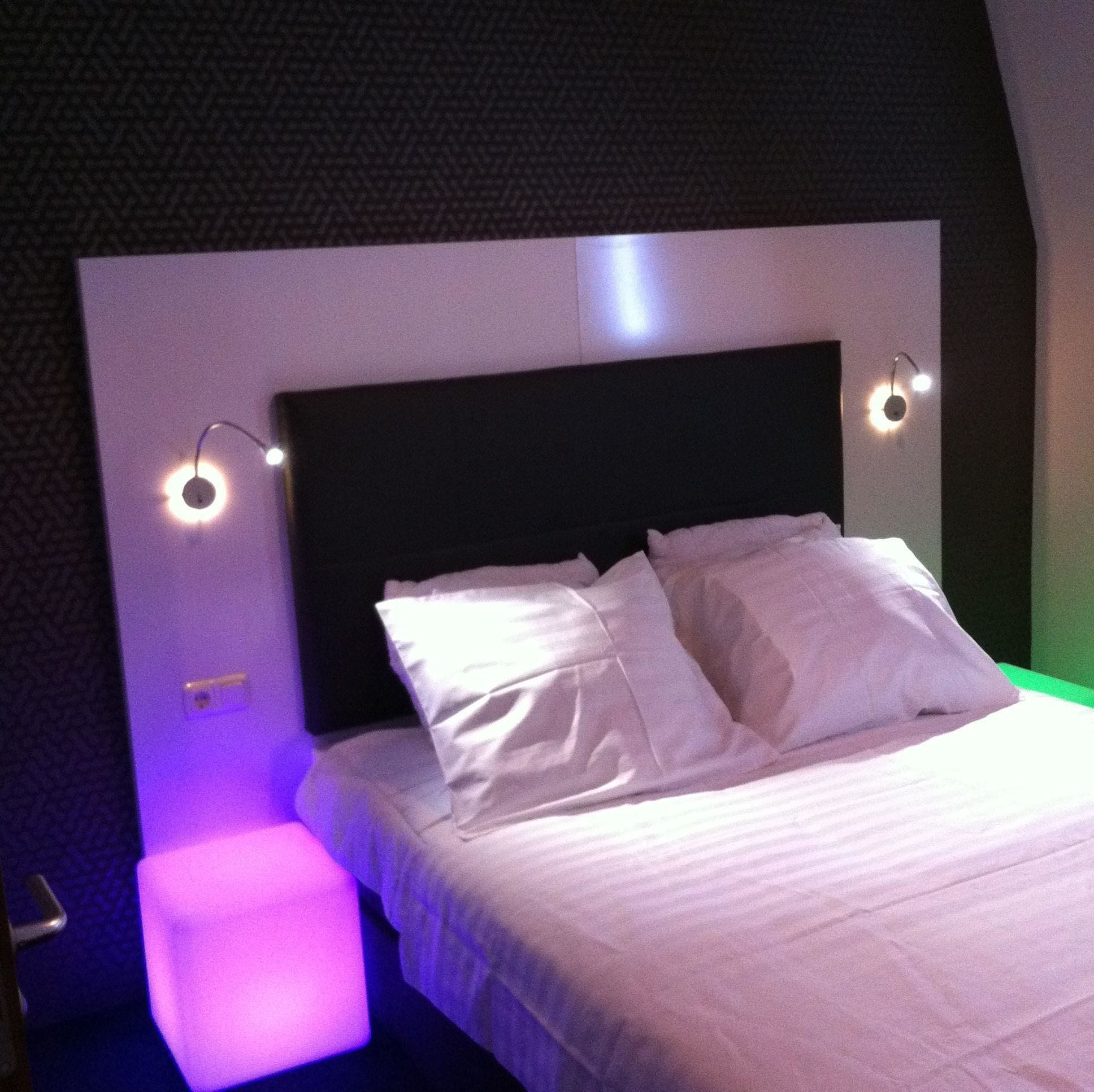 Una cama bien hecha con sábanas y almohadas blancas, cabecera negra y dos lámparas de lectura fijadas a la pared.