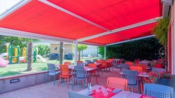 An outdoor patio with tables and chairs, a children's play area in the background, and a red canopy overhead.