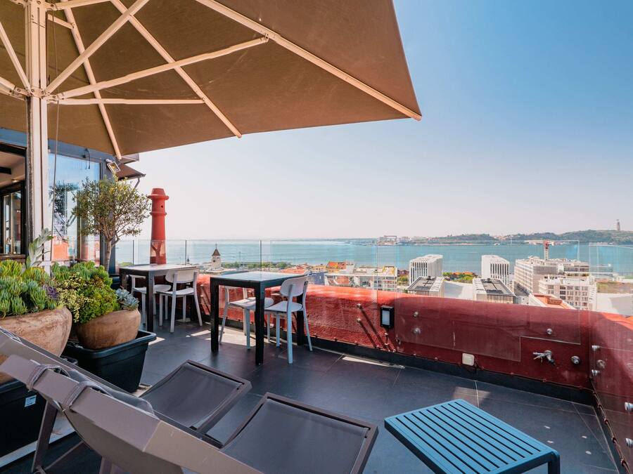 Una terraza en la azotea con mesa y sillas, plantas en macetas y vistas a la ciudad y al mar.