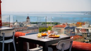 Une table dressée avec des aliments pour le petit-déjeuner et un verre de jus d’orange, donnant sur un paysage urbain avec une tour imposante.
