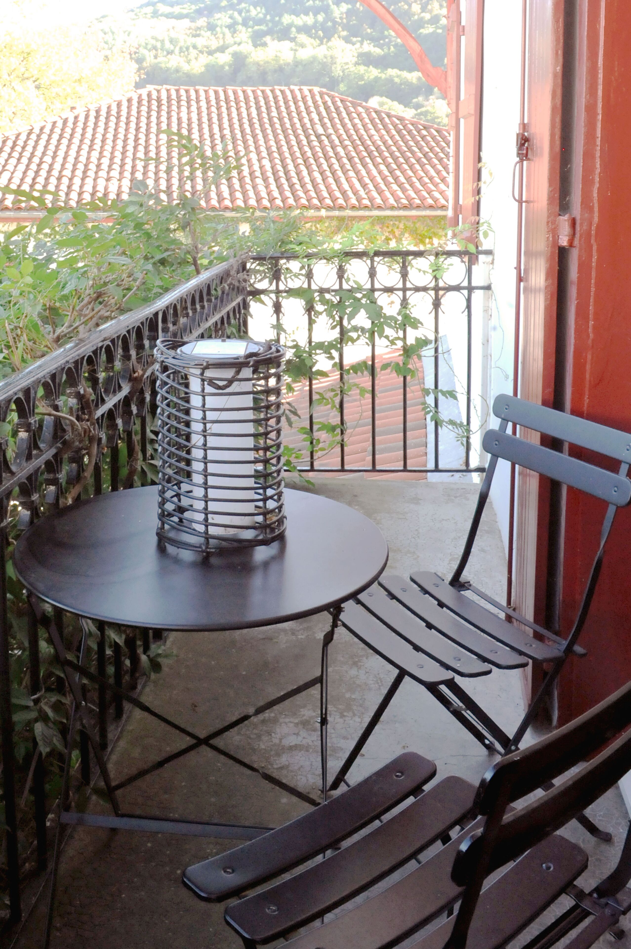 A balcony with a round table and two chairs, a metal lantern, and a view of a red-tiled roof and greenery.